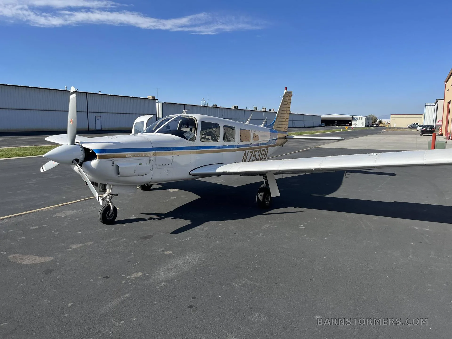 A white and blue Piper Cherokee Lance (N75398) parked on an asphalt ramp with hangars in the background under a clear blue sky.