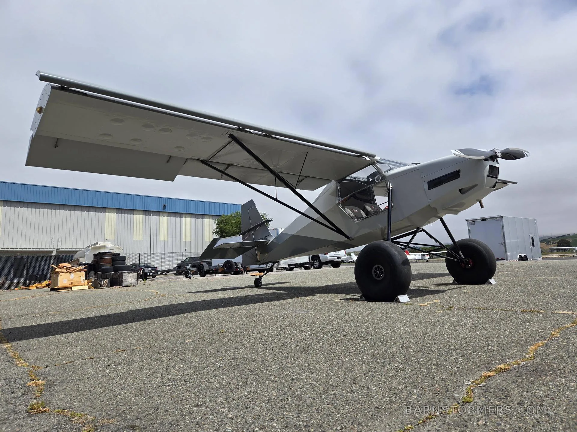 A low-angle view of a light gray high-wing aircraft with large tires parked on an airfield.