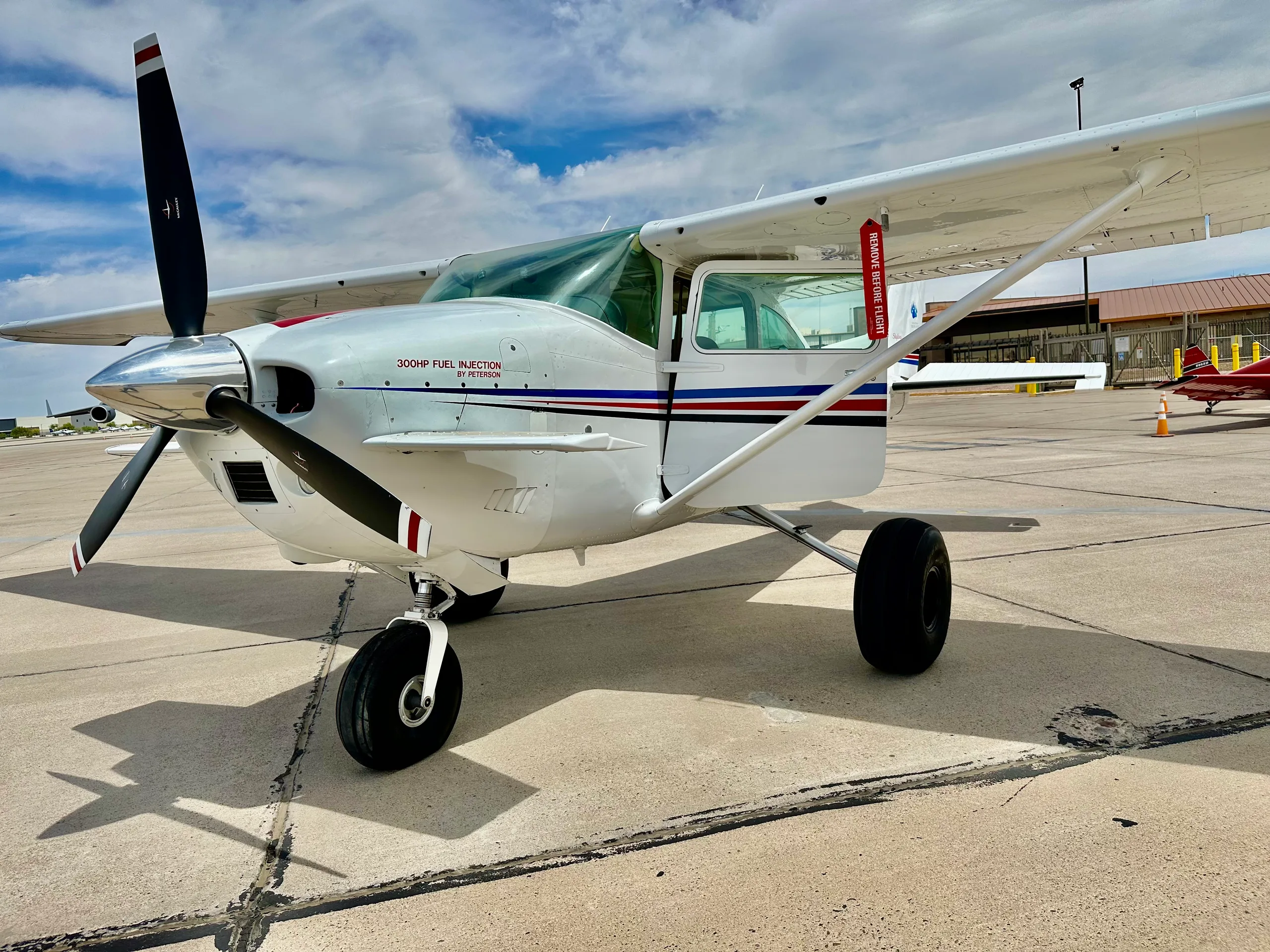 A white single-engine propeller aircraft with red and blue stripes on the fuselage, parked on an asphalt ramp under a partly cloudy sky.