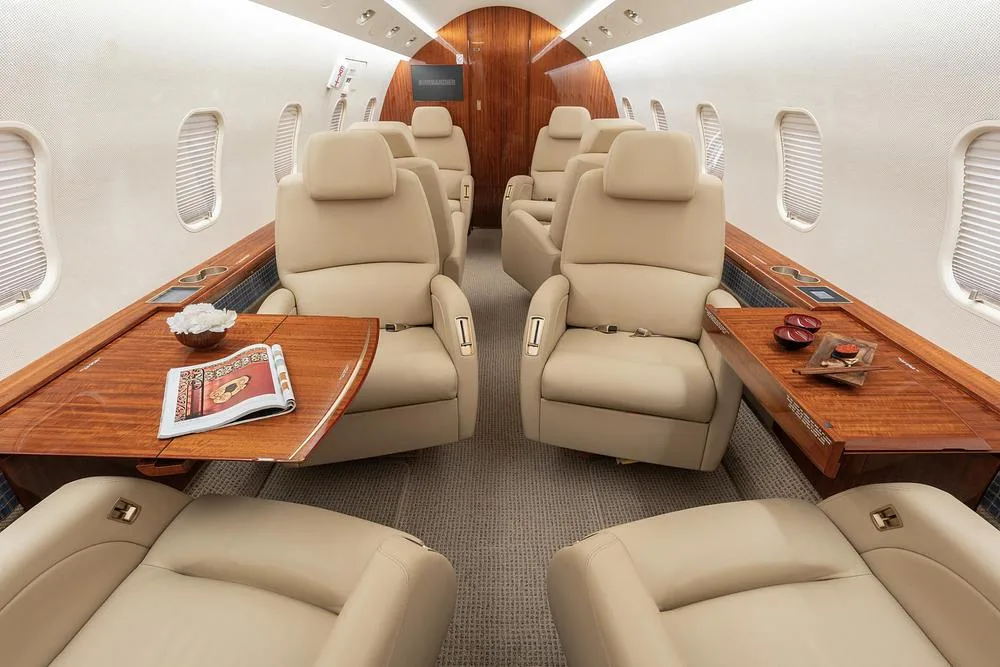 The luxurious interior cabin of a business jet, featuring beige leather seats, polished wood tables, and a rear bulkhead with a "Bombardier" screen.