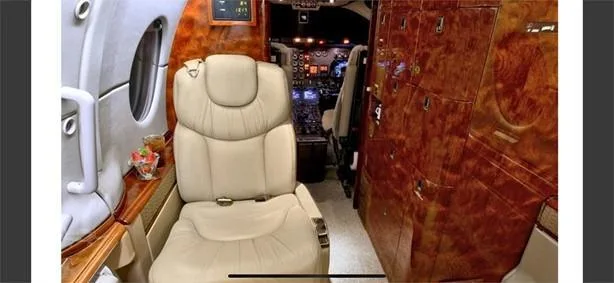 A view from the aircraft cabin showing a beige leather seat, dark wood cabinetry, and a partial view of the cockpit instruments.
