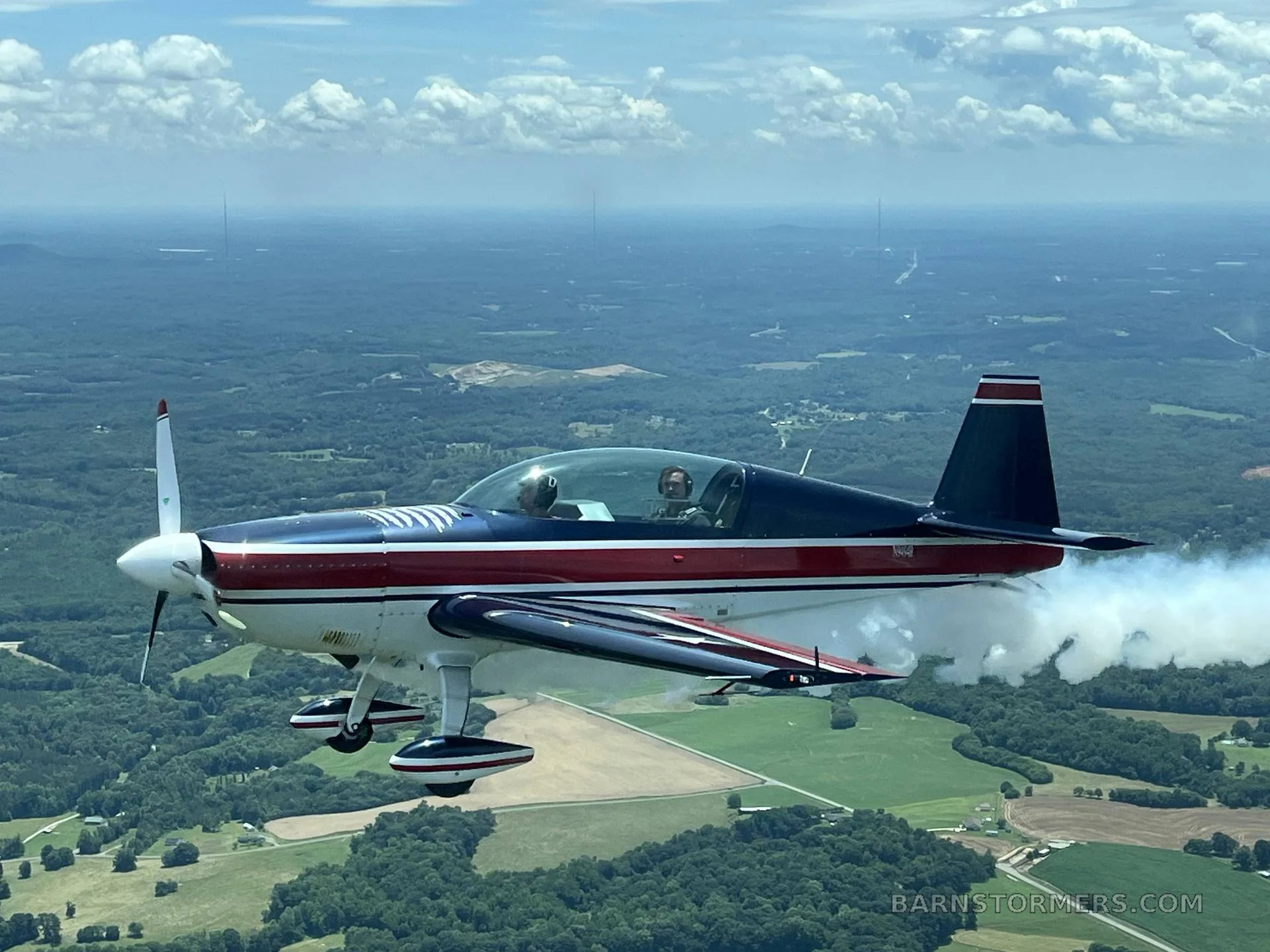 A dark blue, red, and white aerobatic aircraft with stars on its wings, flying level and trailing smoke over a green, hilly landscape.