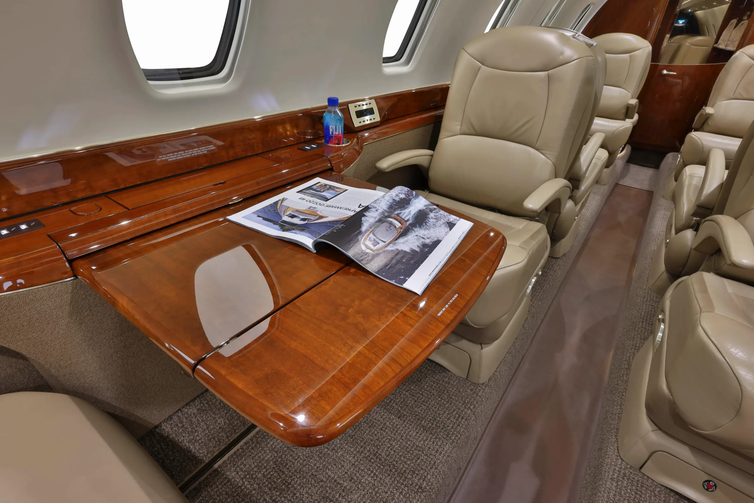 The luxurious interior cabin of a private jet features tan leather seats, a polished wooden table with a magazine, and large windows.