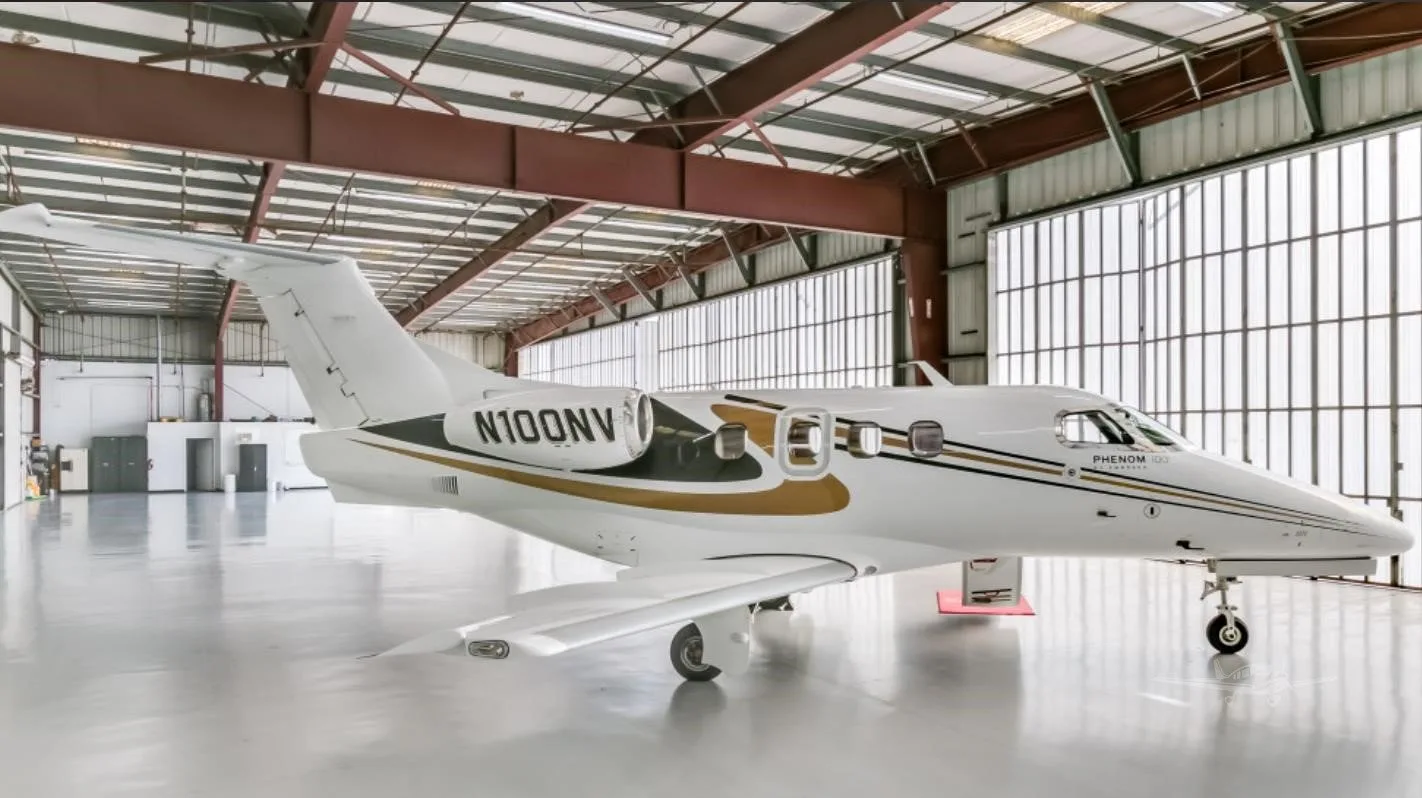 A white Embraer Phenom 100 private jet with gold and black stripes, registration N100NV, parked inside a hangar.