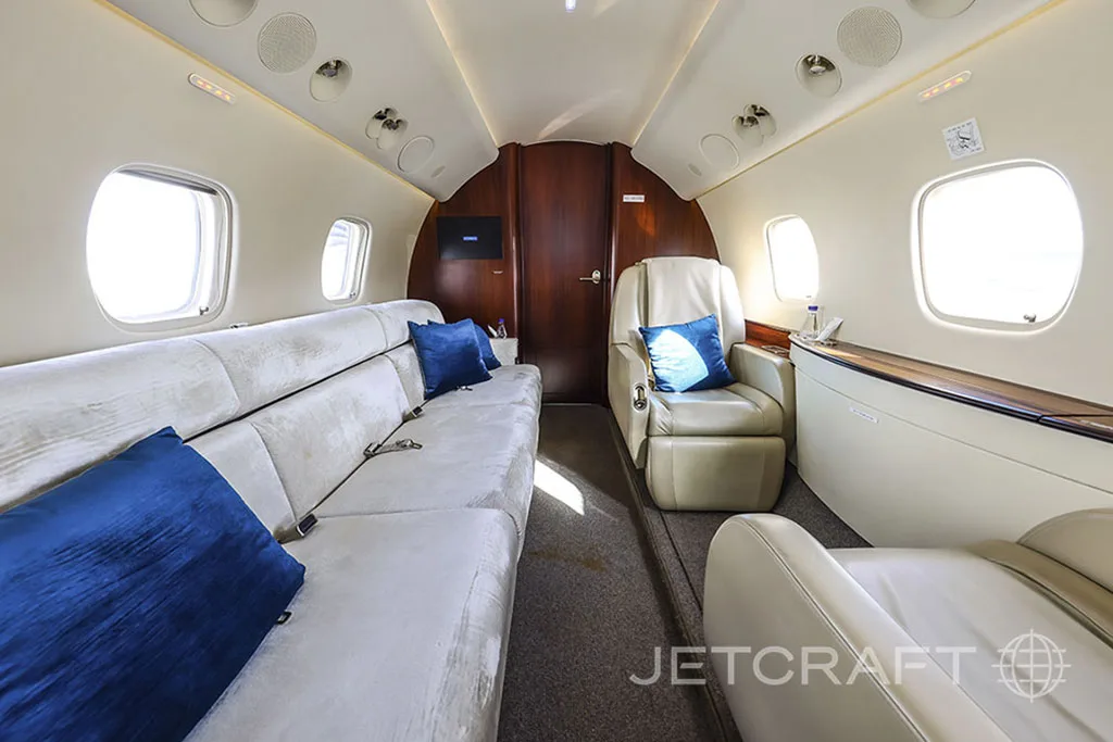 A section of the aircraft cabin with a long light-colored sofa and a single armchair, both accented with blue pillows.