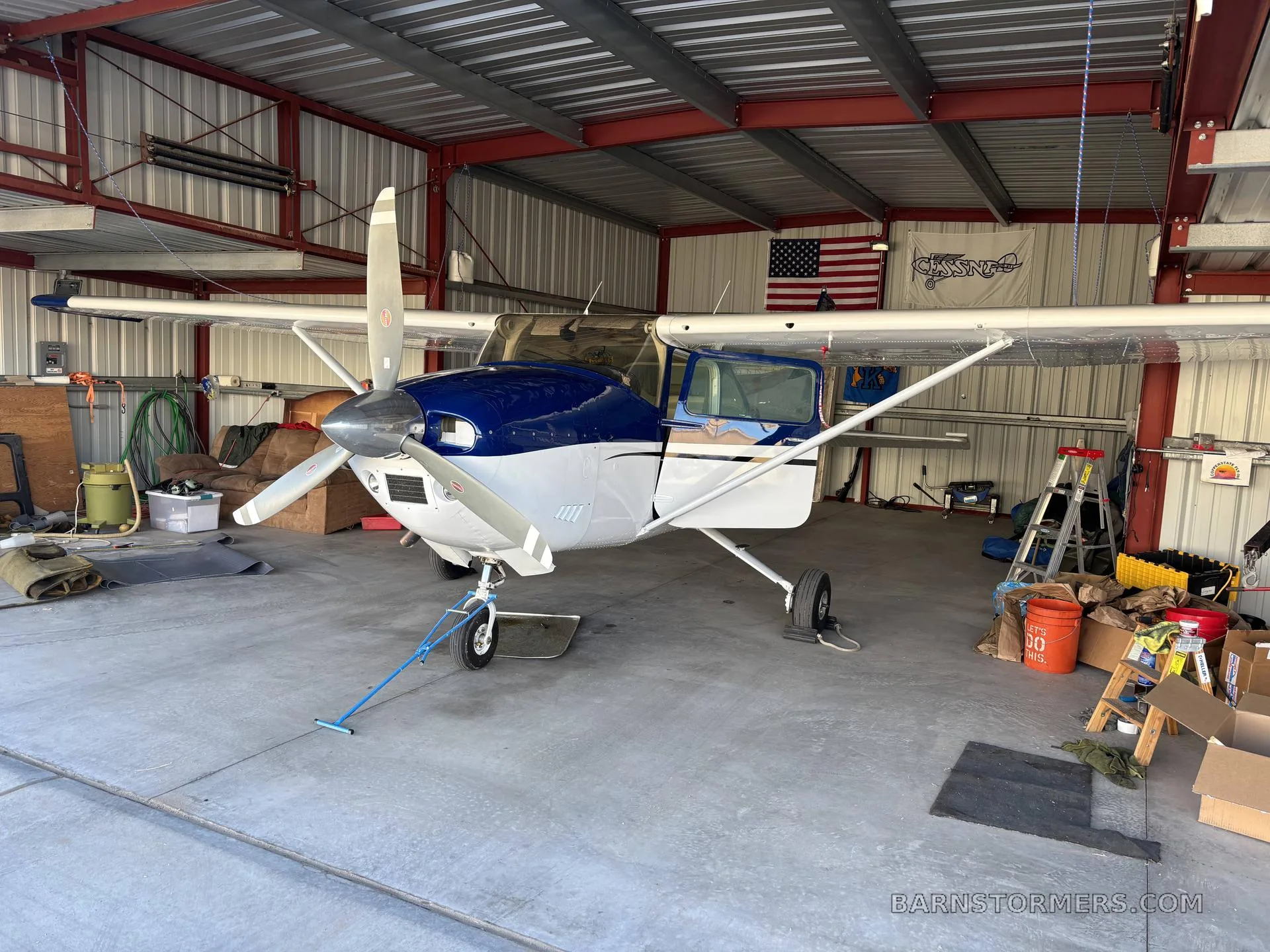 A blue and white single-engine propeller aircraft is parked inside a metal hangar with its door open.