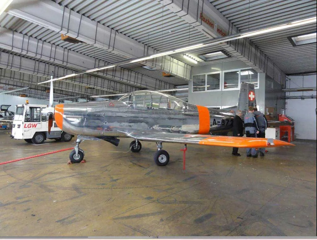 A side view of a polished silver and orange Pilatus P-3 aircraft with Swiss markings, parked inside a hangar.