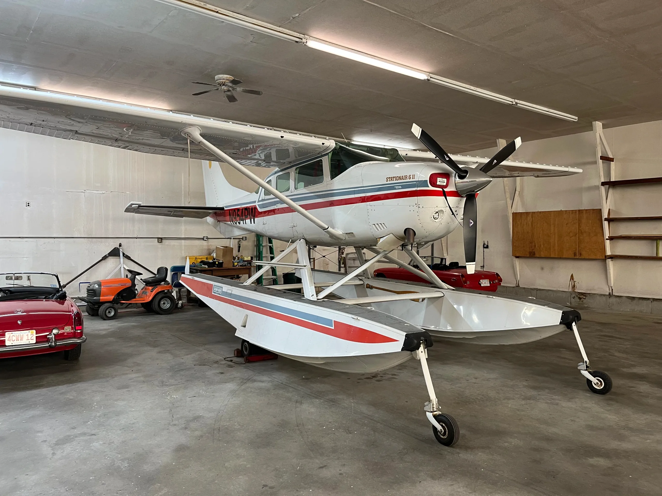 A white and red Cessna Stationair 6 II floatplane with registration N954PW is parked in a hangar, with a red convertible car and a lawnmower nearby.