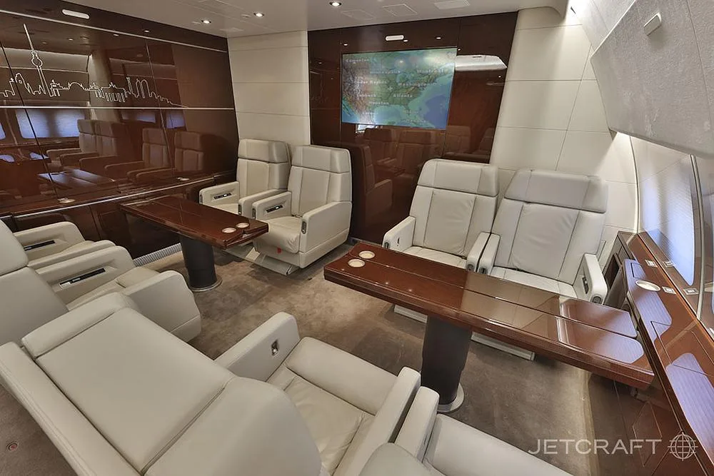 A luxurious aircraft interior with cream leather lounge chairs, dark wood tables, and a wall-mounted map display.