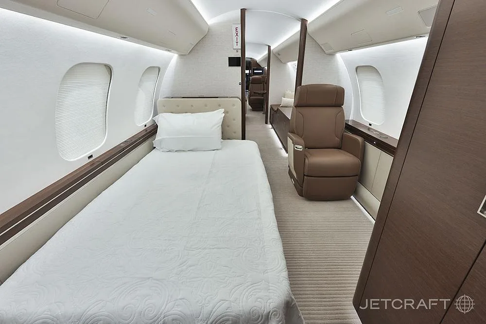 A business jet cabin interior is configured with a bed made up with white bedding and a brown leather seat.