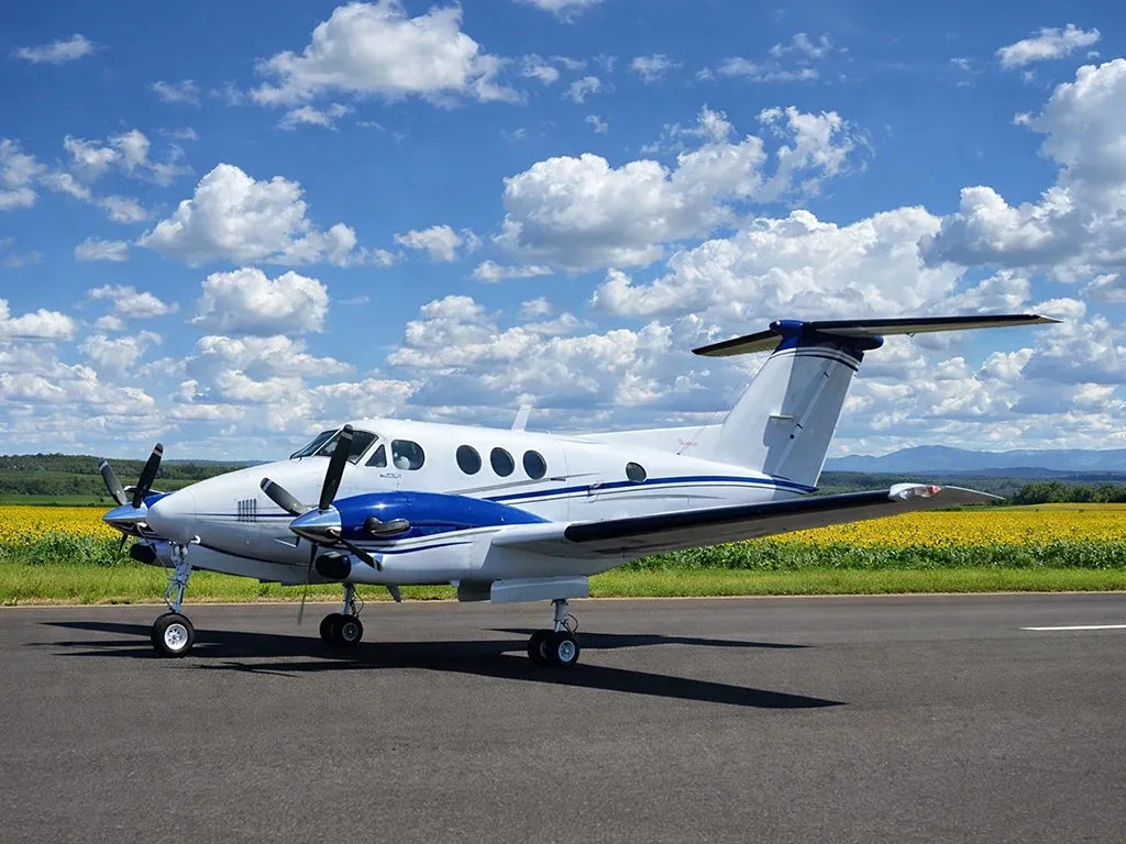 A white and blue Beechcraft King Air turboprop aircraft is parked on an airfield with a field of sunflowers and mountains in the background under a blue sky.