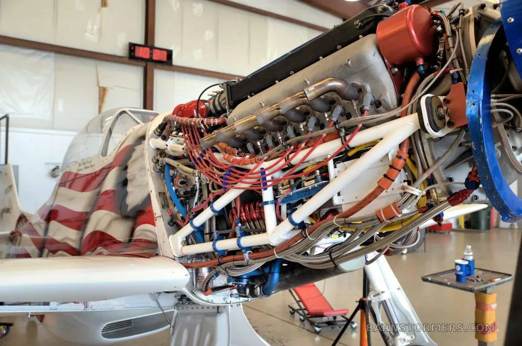 The exposed engine and wiring of an American flag-painted aircraft, likely a P-51 Mustang replica, in a hangar.