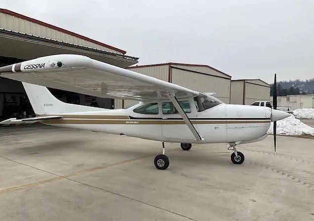 A side view of the white Cessna 172 (N7280C) with gold and black stripe, parked on a tarmac next to hangars.