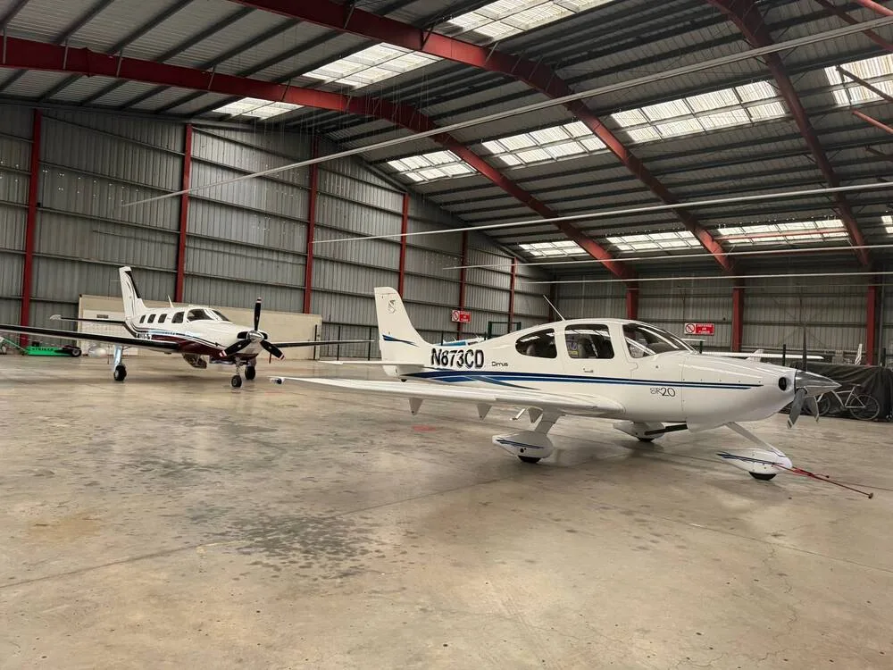 A white Cirrus SR20 aircraft (N873CD) with blue stripes parked inside a hangar next to another twin-engine aircraft.