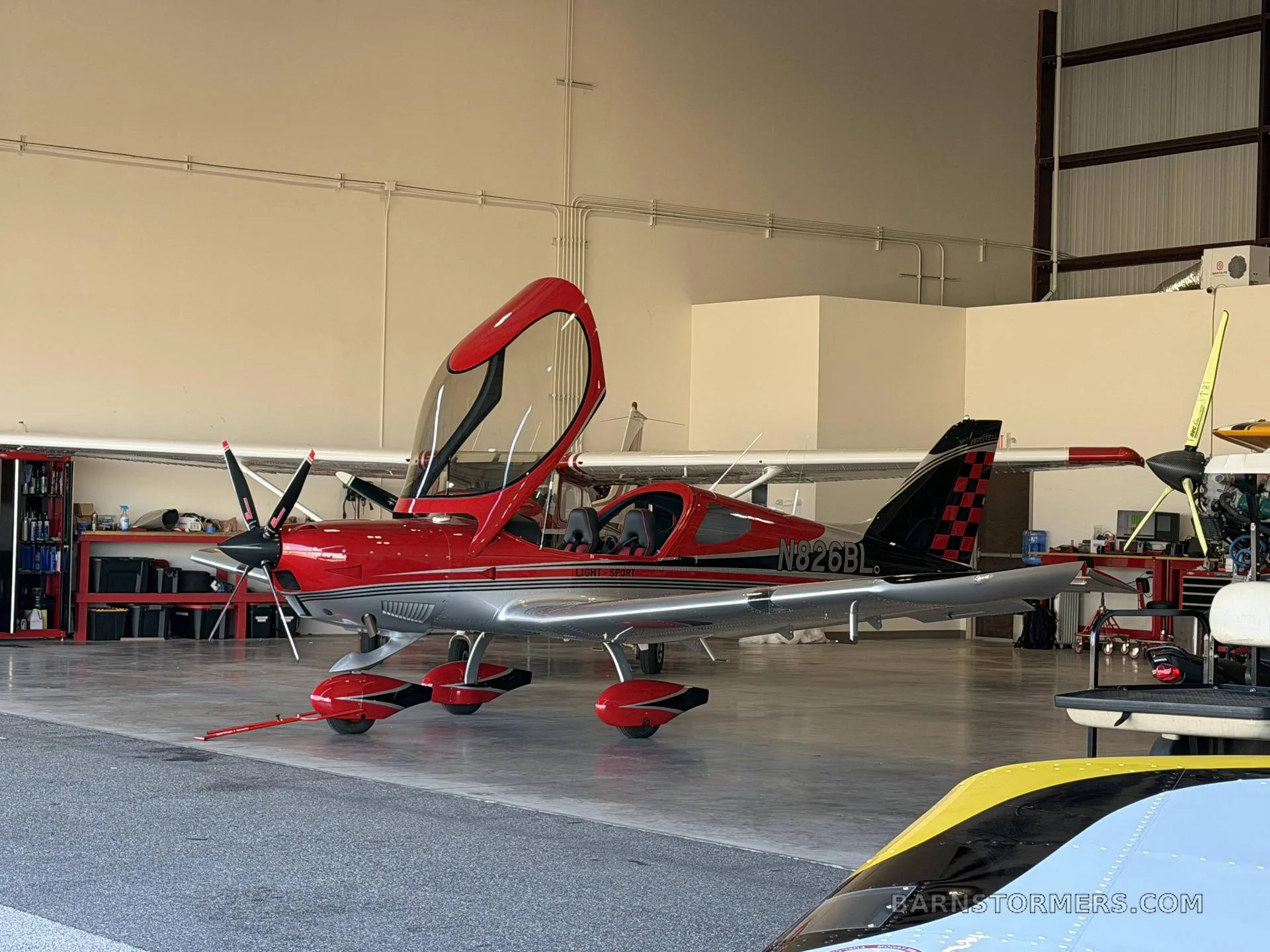 A red and silver Bristell LSA aircraft with its canopy open, parked in a hangar.