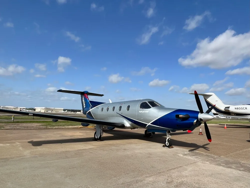 A Pilatus PC-12 aircraft with a silver and blue paint scheme parked on an airfield under a partly cloudy sky.