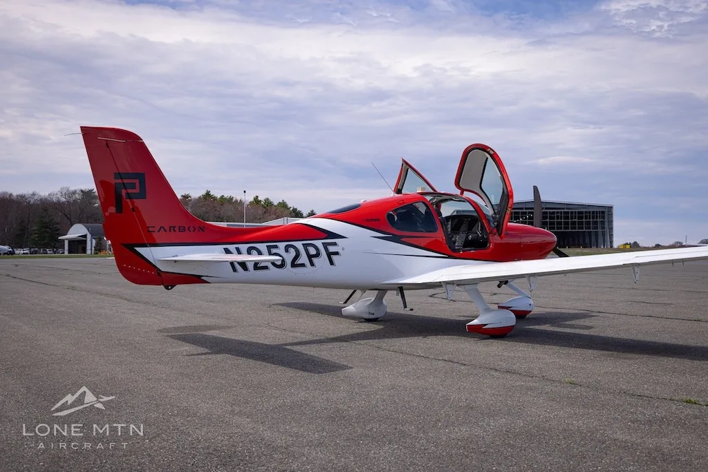 A rear-side view of a red and white Cirrus SR20 aircraft with its doors open, parked on an airfield.