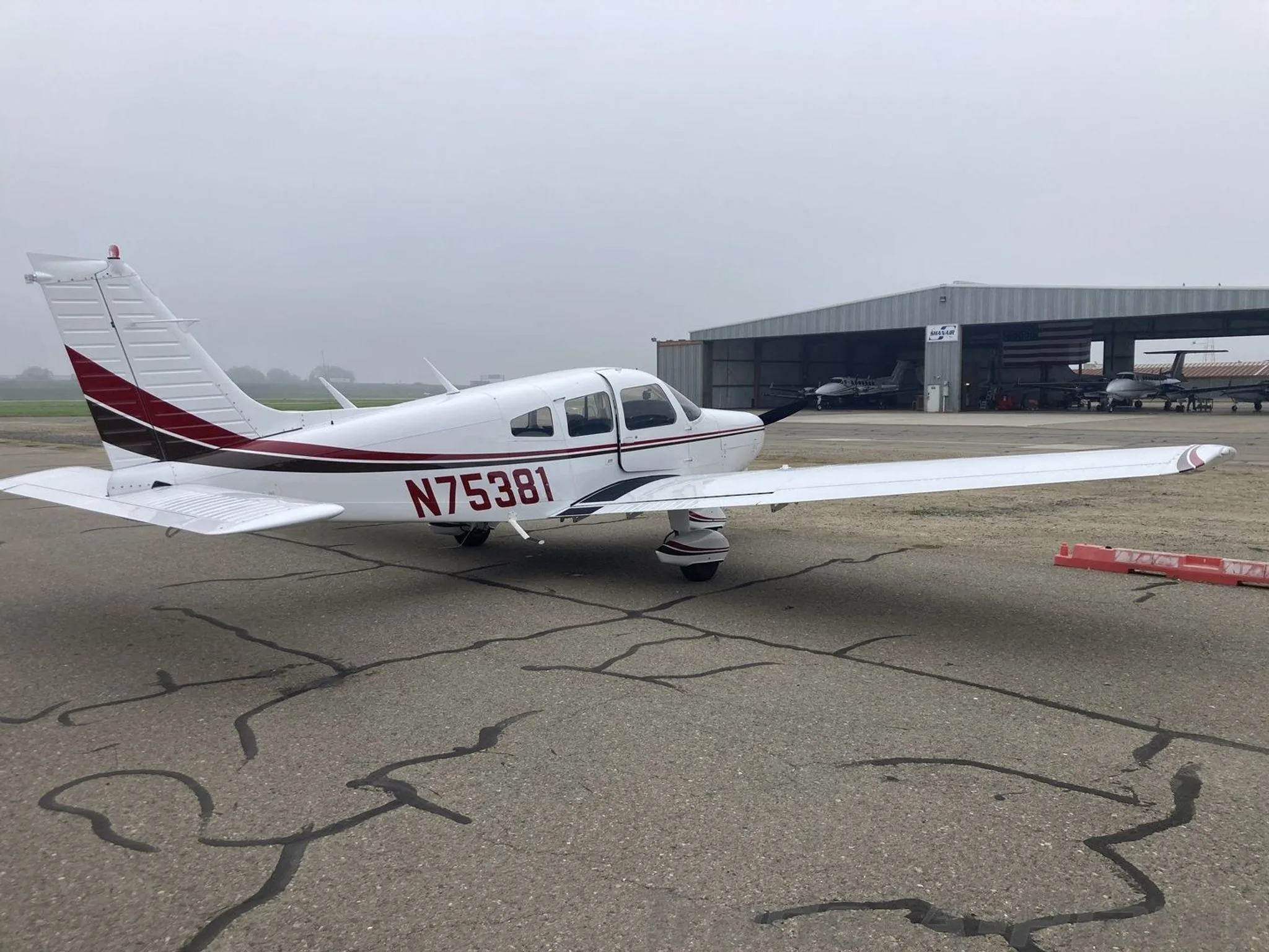 A white Piper PA-28-151 aircraft with red and black stripes parked on an airfield with hangars in the background.