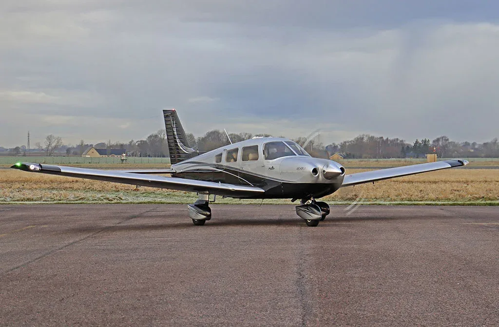 A silver and black Piper Archer LX aircraft is parked on an airfield under a cloudy sky.