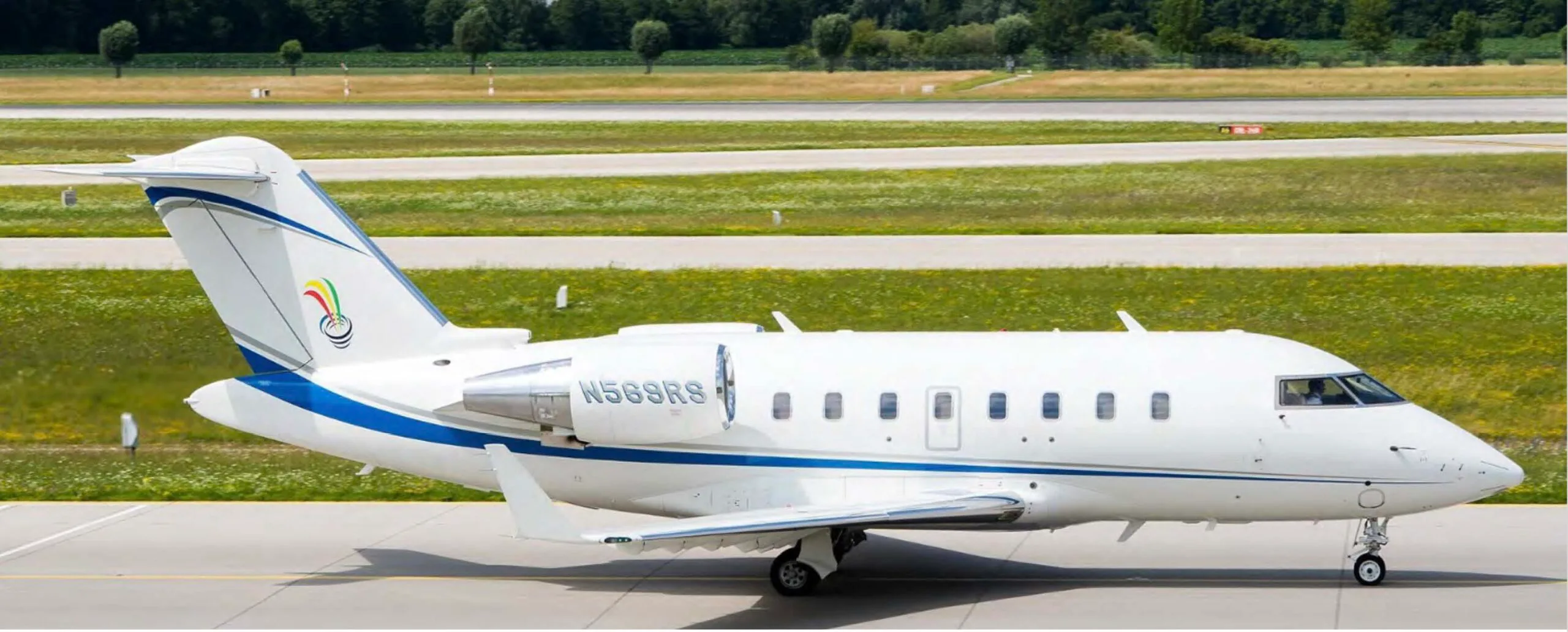 A white Bombardier Challenger business jet with blue and grey stripes and a tail logo, parked on a tarmac.