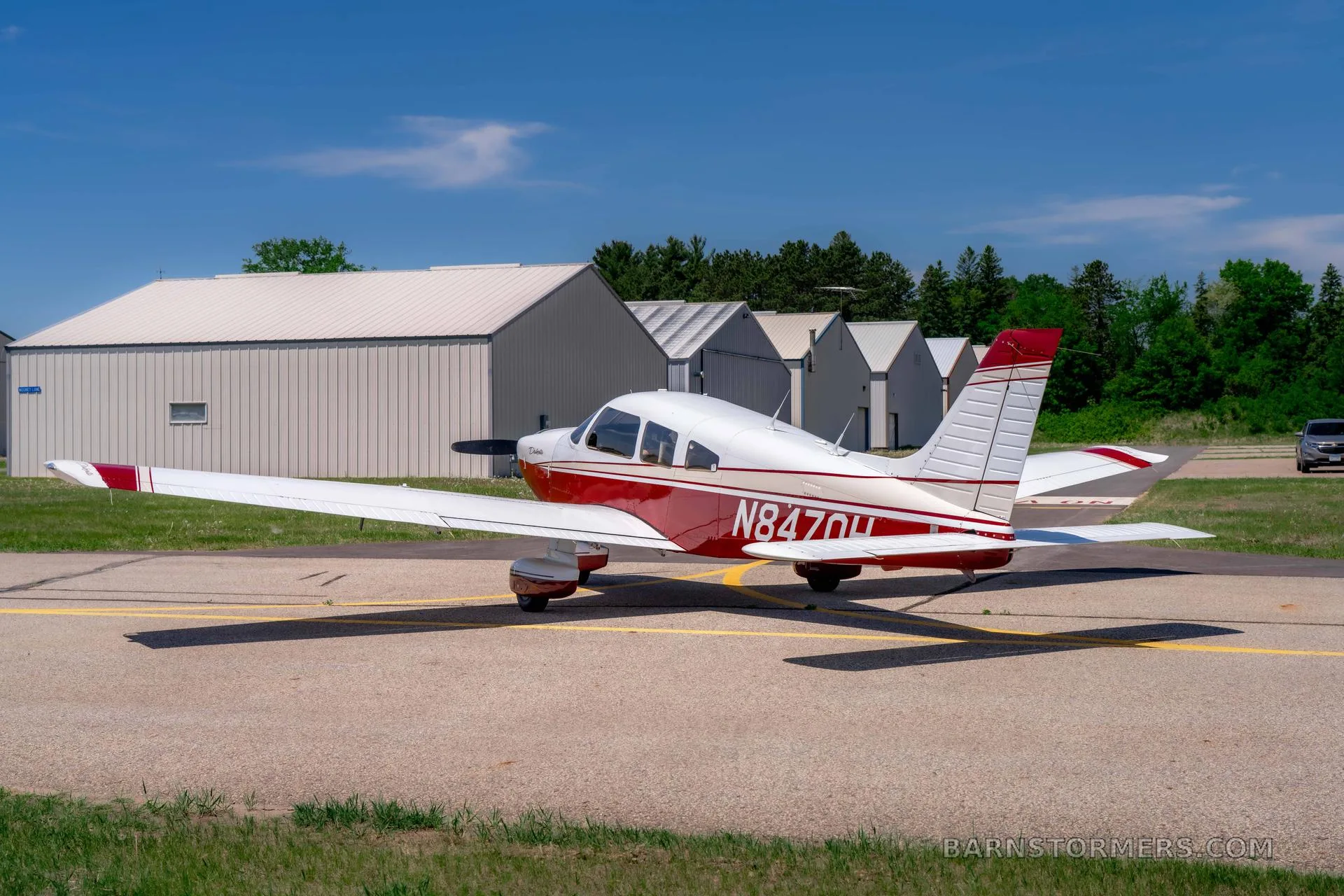 A red and white Piper Dakota aircraft with registration N847OH is parked on an asphalt tarmac, viewed from the rear quarter, with hangars in the distance.