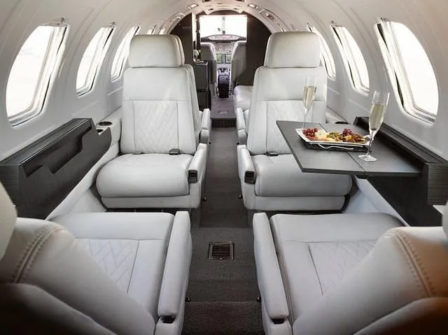 A view down the aisle of a private jet cabin, showing multiple white leather seats and a table set with champagne and snacks.