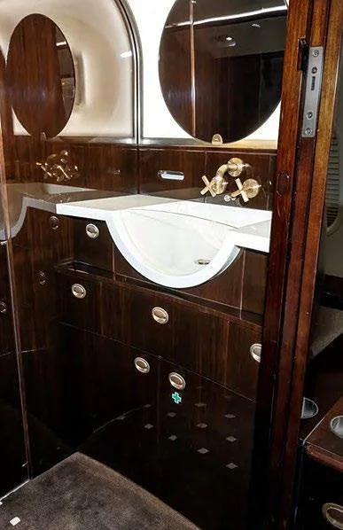 A luxurious aircraft lavatory with a white sink, gold fixtures, and dark polished wood cabinetry and mirrors.