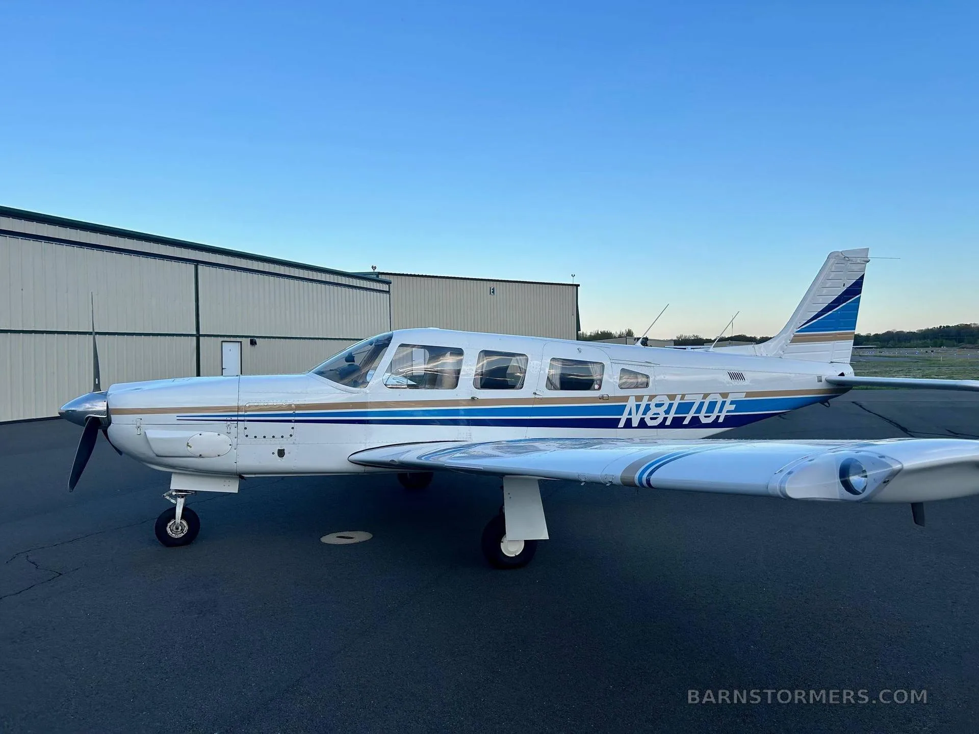 A white single-engine propeller aircraft with blue and gold stripes and tail number N8170F, viewed from the left side, parked next to a hangar under a clear blue sky.