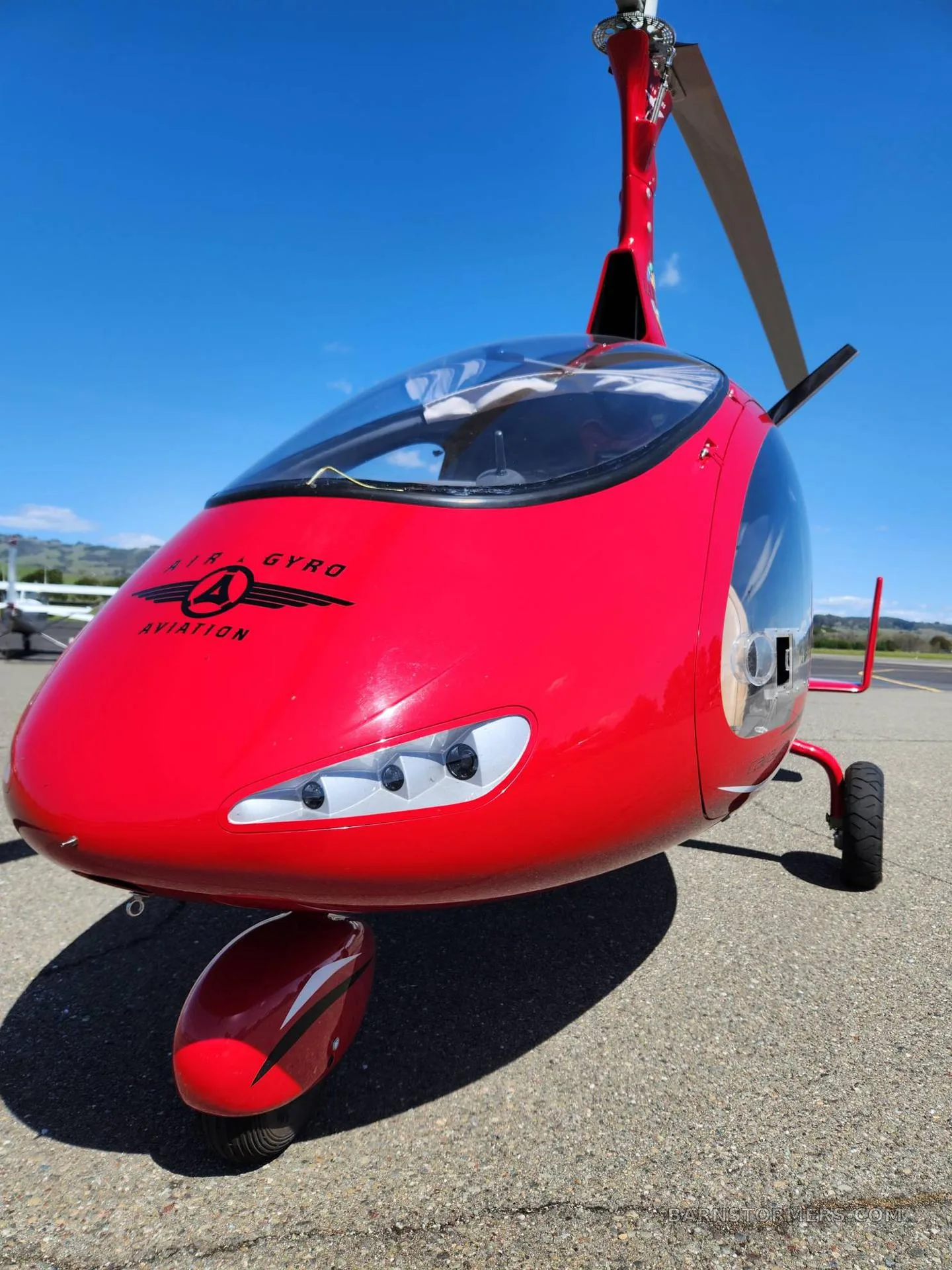 A front view of the red gyrocopter, featuring the 'AIR GYRO AVIATION' logo on its nose.
