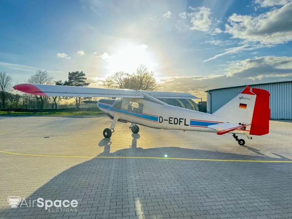 A white aircraft with blue and red stripes and red wingtips, parked on a paved apron with the sun in the background.