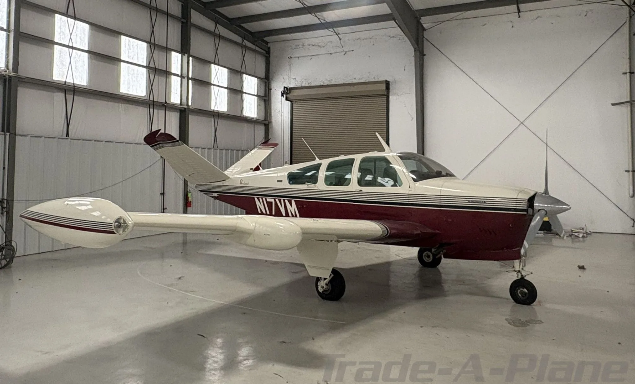 A white and maroon Beechcraft Bonanza N17VM parked inside a hangar, viewed from the rear-left.