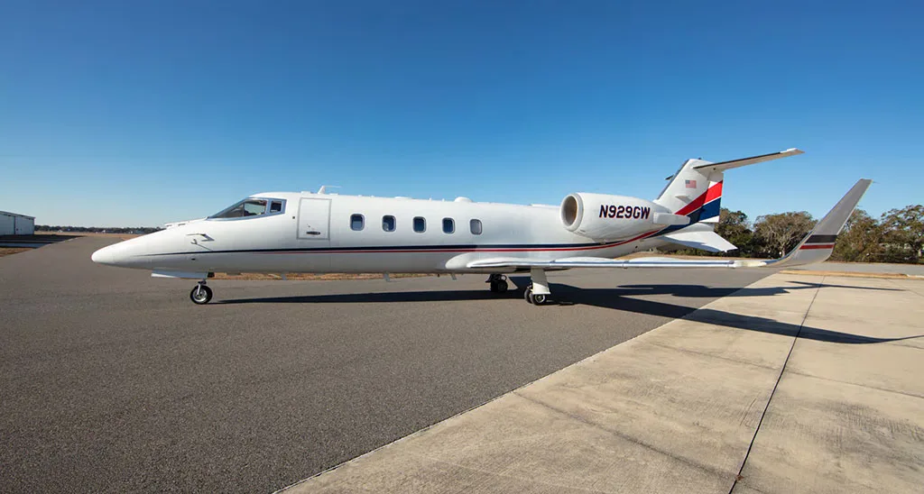 A white private jet with red and blue tail stripes, registration N929GW, parked on an asphalt and concrete apron under a clear blue sky.