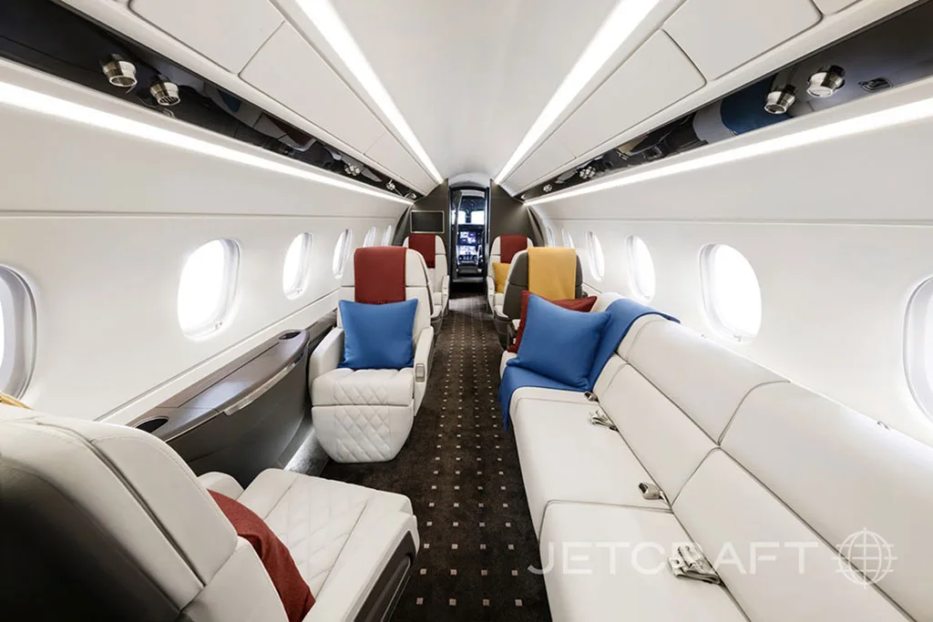 The spacious interior cabin of a business jet featuring white quilted seating, a sofa, and blue and red accent pillows.