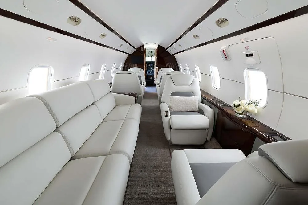The spacious interior cabin of a business jet, showing a long white leather sofa and individual white and grey seats.