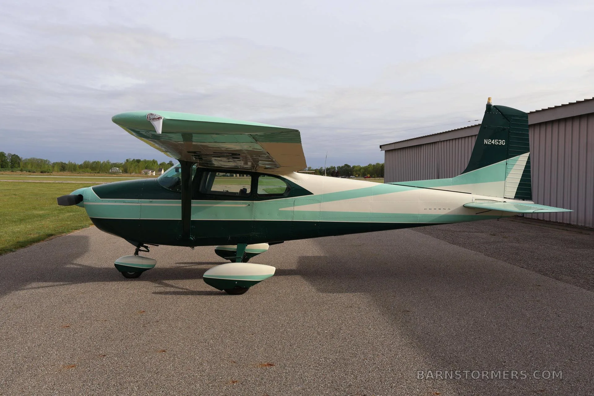 A green and white single-engine propeller aircraft (Cessna 172) is parked on an asphalt ramp next to a hangar, viewed from the side.