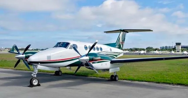 A white turboprop aircraft with green and red stripes is parked on an airport ramp, with an air traffic control tower in the background.