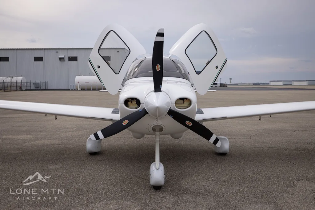 A head-on view of a white Cirrus SR22 aircraft with its propeller and open doors visible, parked on an asphalt ramp.