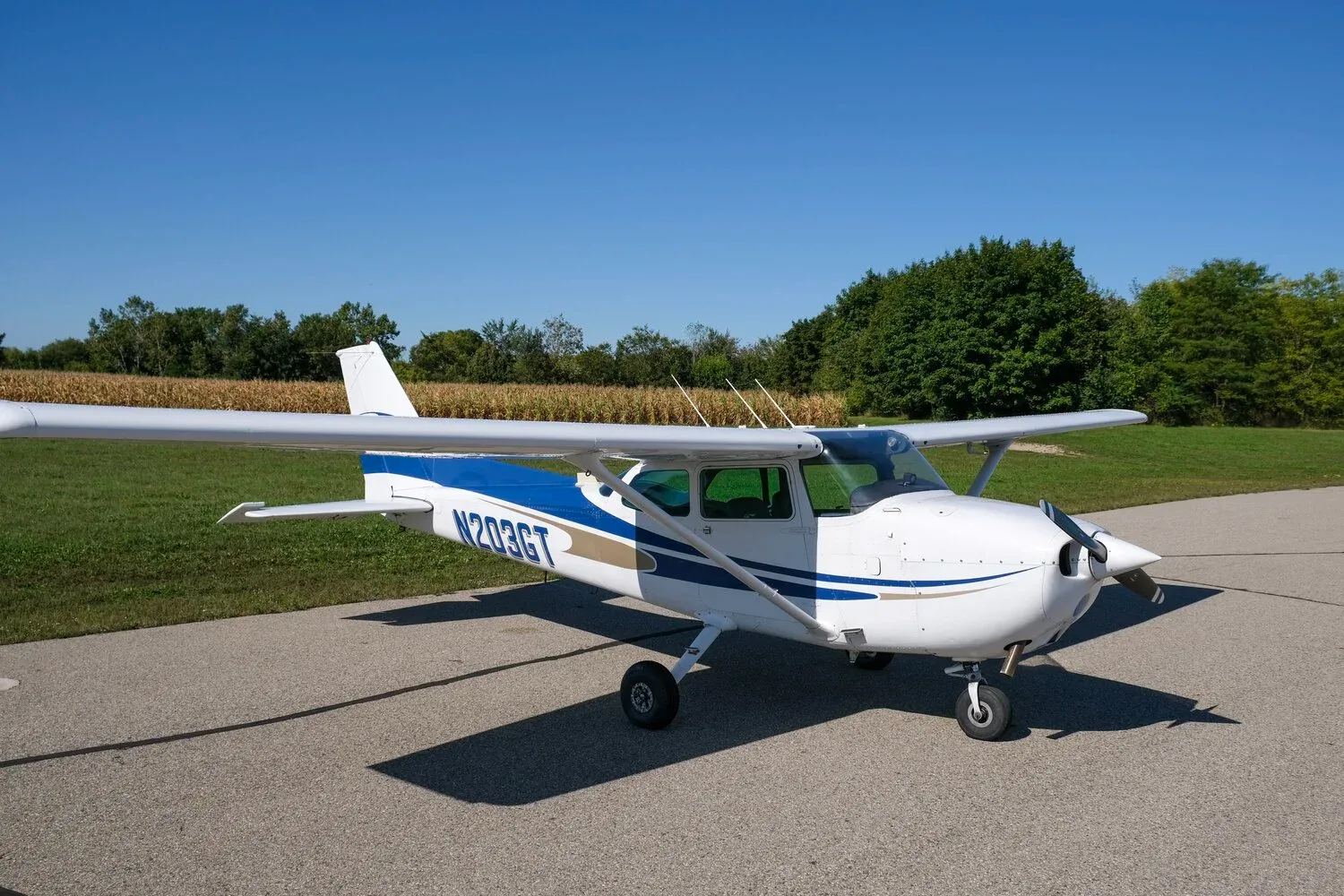 A white Cessna 172 with blue and gold stripes is parked on an airfield under a clear blue sky.