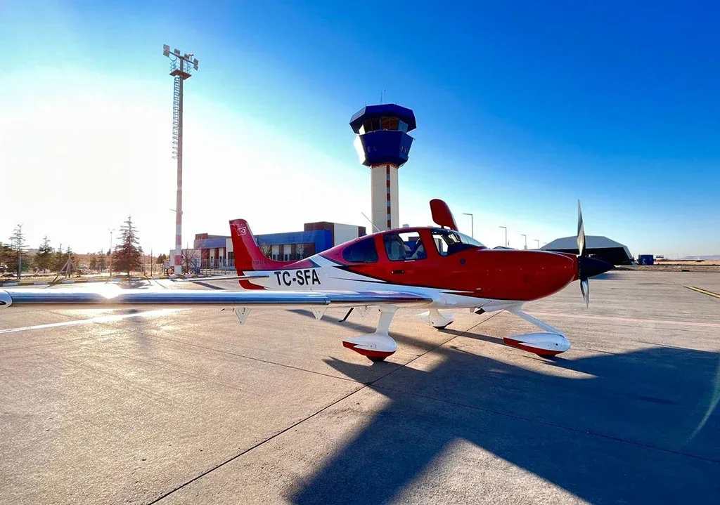 A red and white single-engine aircraft (TC-SFA) parked on an airfield with a control tower under a clear blue sky.