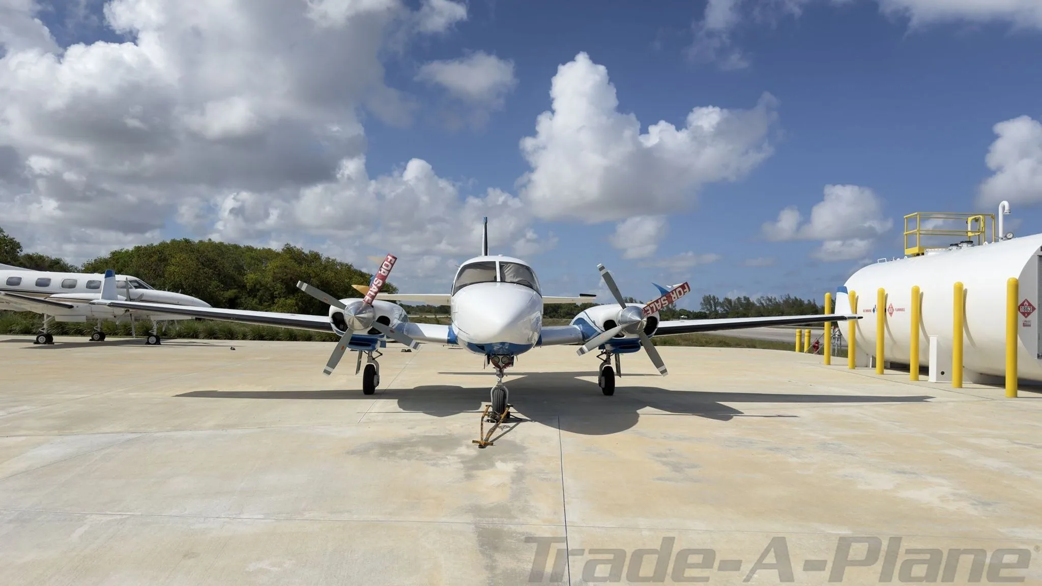 Front view of a white and blue twin-engine propeller aircraft, N348GG, with 'FOR SALE' tags, parked on a concrete ramp next to a fuel tank.