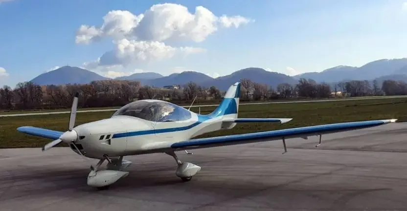 A white and blue light aircraft with a propeller is parked on an asphalt runway with mountains in the background.