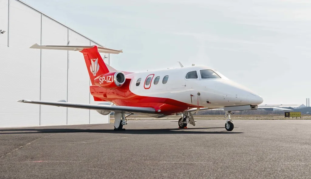 A white and red Embraer Phenom 300, registration SP-IZI, parked on an airport tarmac next to a hangar.