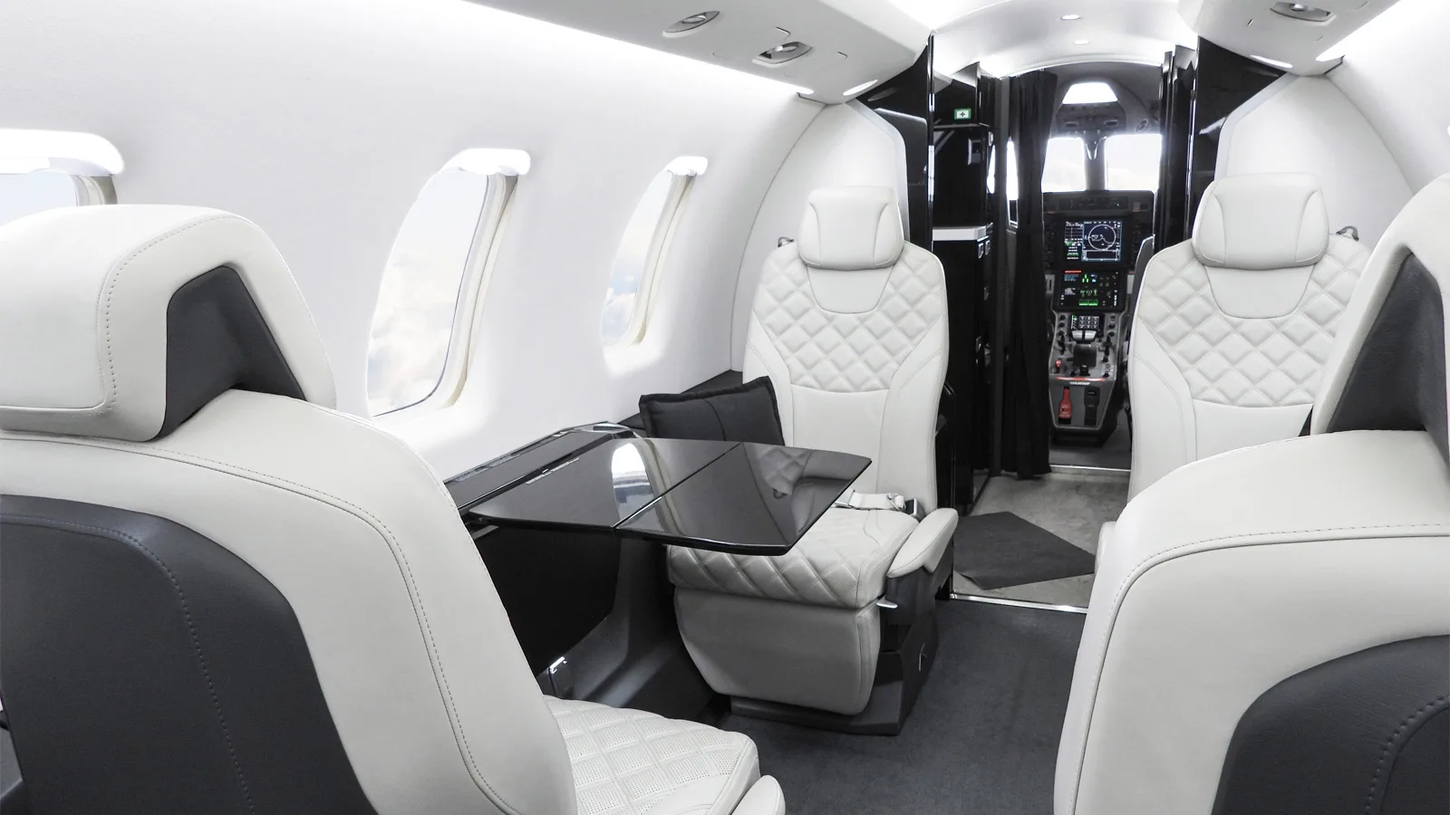 The luxurious white and dark grey interior cabin of a business jet, featuring quilted seats and a fold-out table.