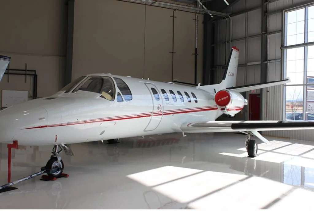 A white Cessna Citation aircraft with red stripes parked in a hangar, viewed from the front-left.