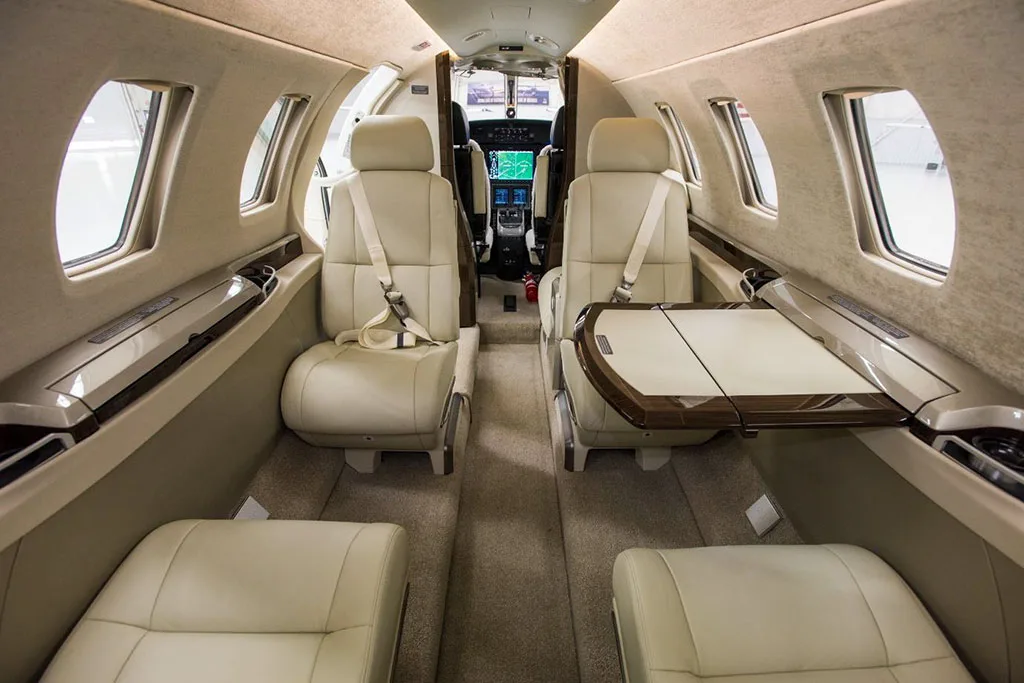 The beige interior cabin of a private jet, showing passenger seats and a view into the cockpit.