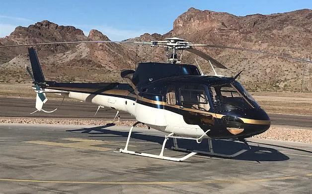 A dark blue and white helicopter with gold stripes is parked on an airfield with mountains in the background.