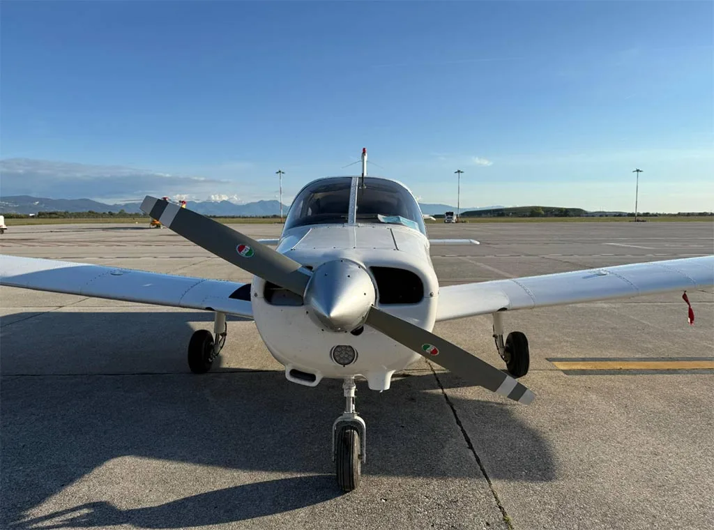 A head-on view of a white single-engine propeller aircraft with a two-bladed propeller, parked on an airfield.