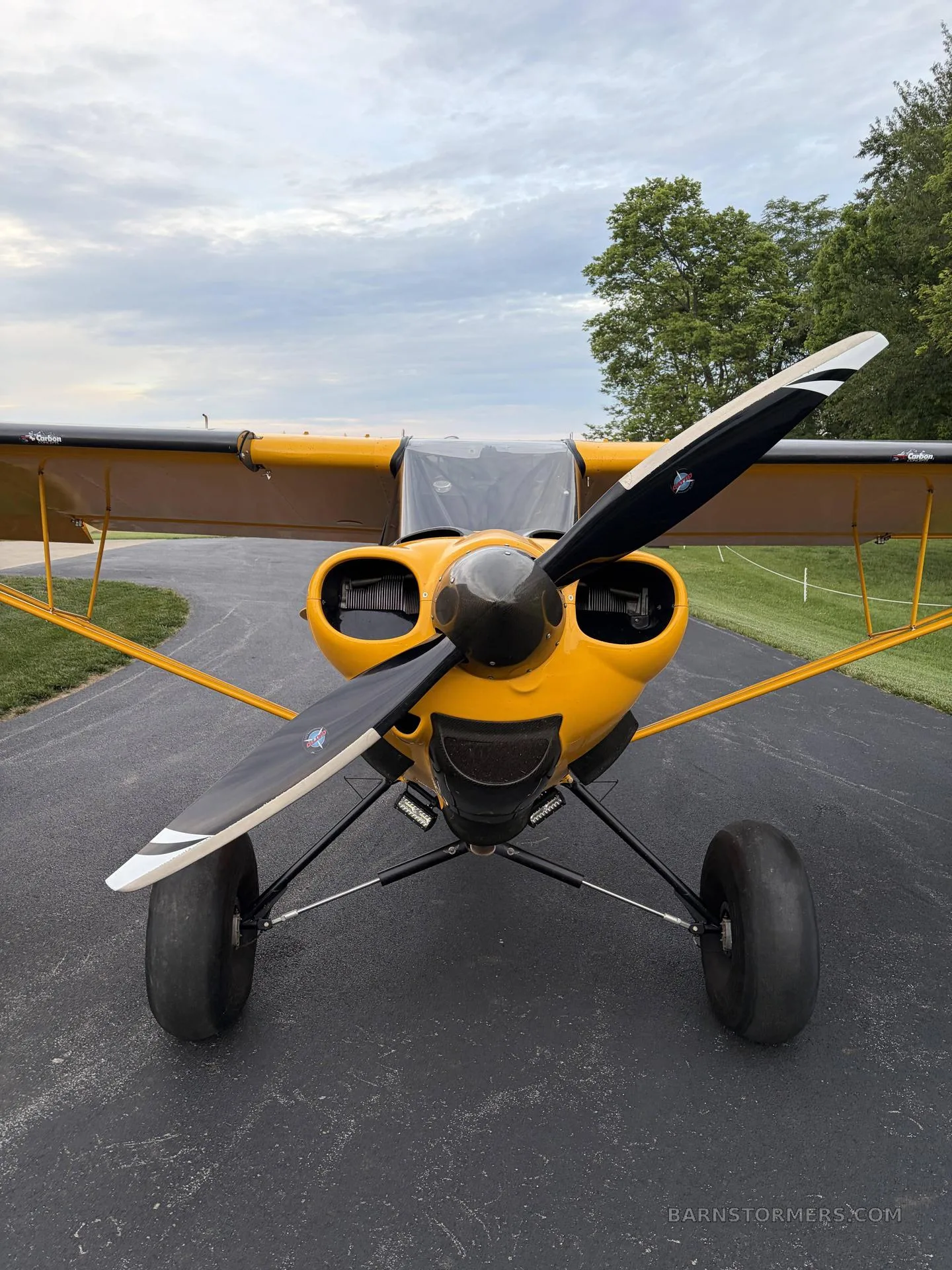 A head-on view of a yellow Carbon Cub aircraft with a black propeller and large tundra tires on an asphalt surface.