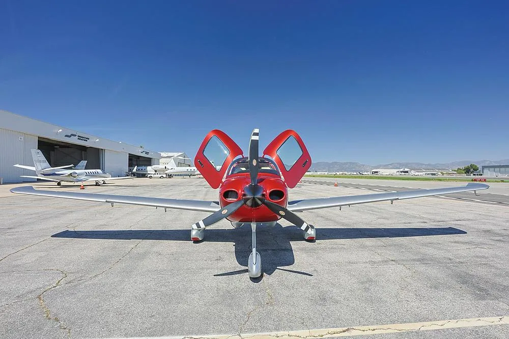 A front view of a red and silver Cirrus SR22 aircraft with registration N218JS and open doors on an airport tarmac.