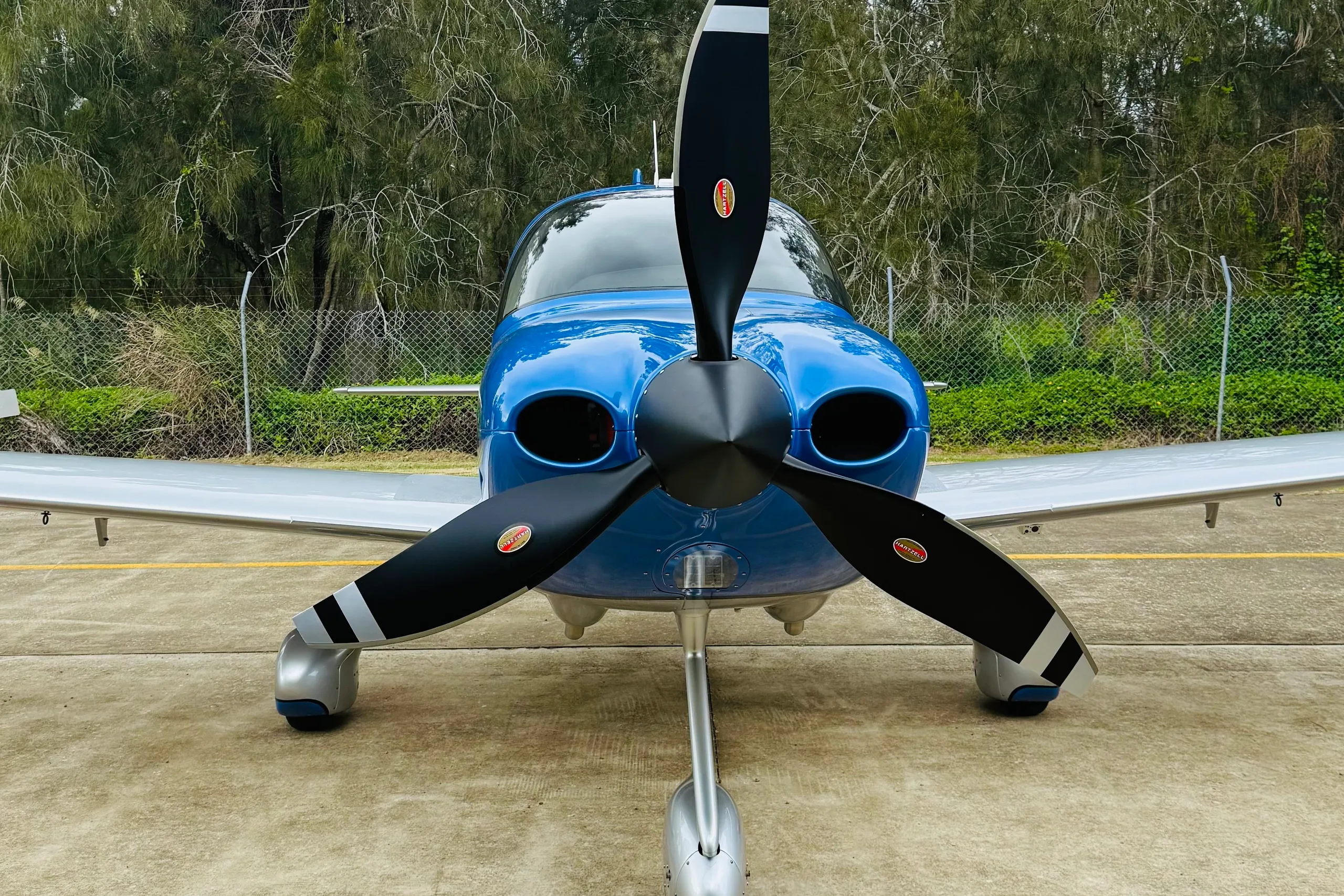 A front view of a blue and silver single-engine propeller aircraft with a black Hartzell propeller, parked on a concrete tarmac.